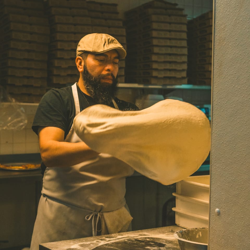 A pizza chef in a flat cap and apron tosses pizza dough in the air.