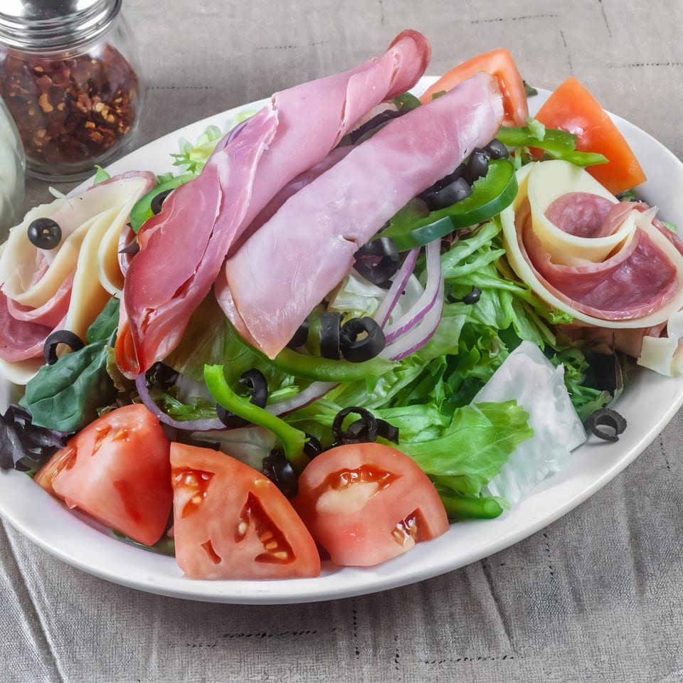 A large Italian salad with ham, salami, cheese, olives, tomatoes, and peppers on a white plate.