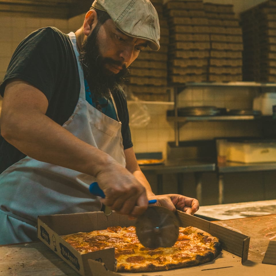A pizzaiolo in a cap and apron slices a pepperoni pizza in a box at Luisa's.