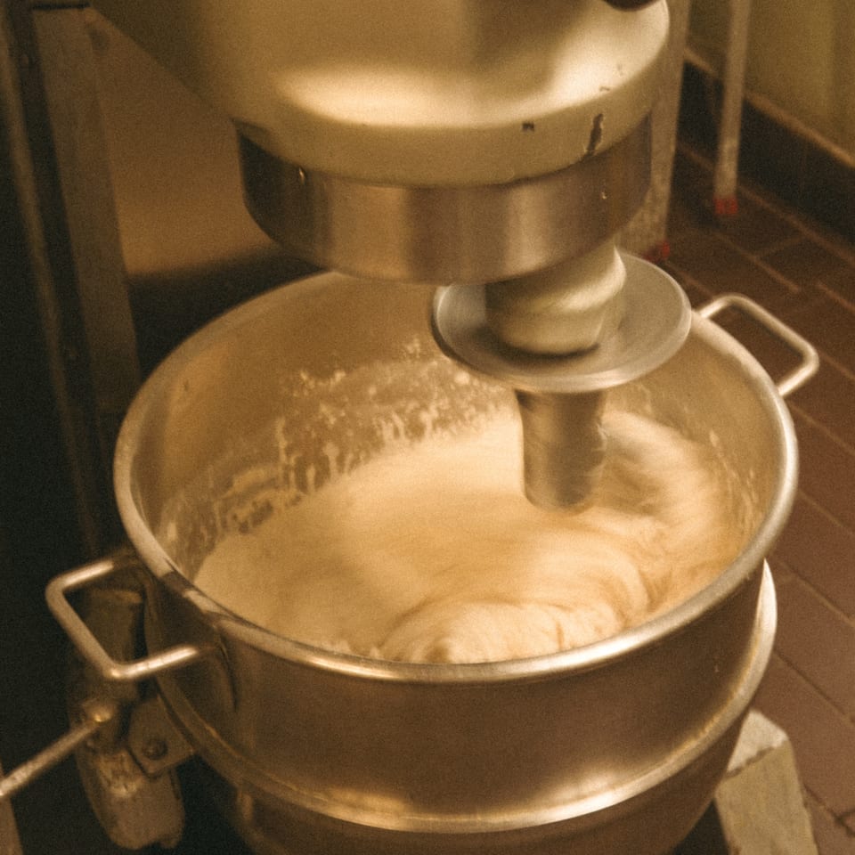 A commercial mixer vigorously kneads pizza dough in a large metal bowl at Luisa's Pizzeria.