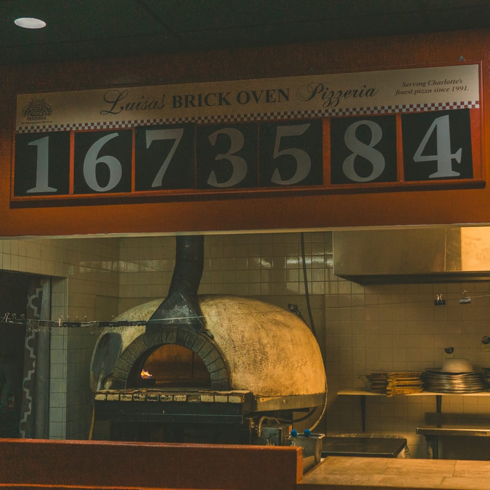 Luisa's Brick Oven Pizzeria sign above a large, domed brick oven with glowing embers inside.
