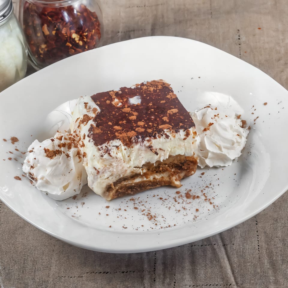 A slice of tiramisu with whipped cream and cocoa powder on a white plate.